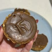 Foreground of image is a close up of a caramel tart being held up for the camera, background is caramel tarts sitting on a blue plate slightly blurred out.
