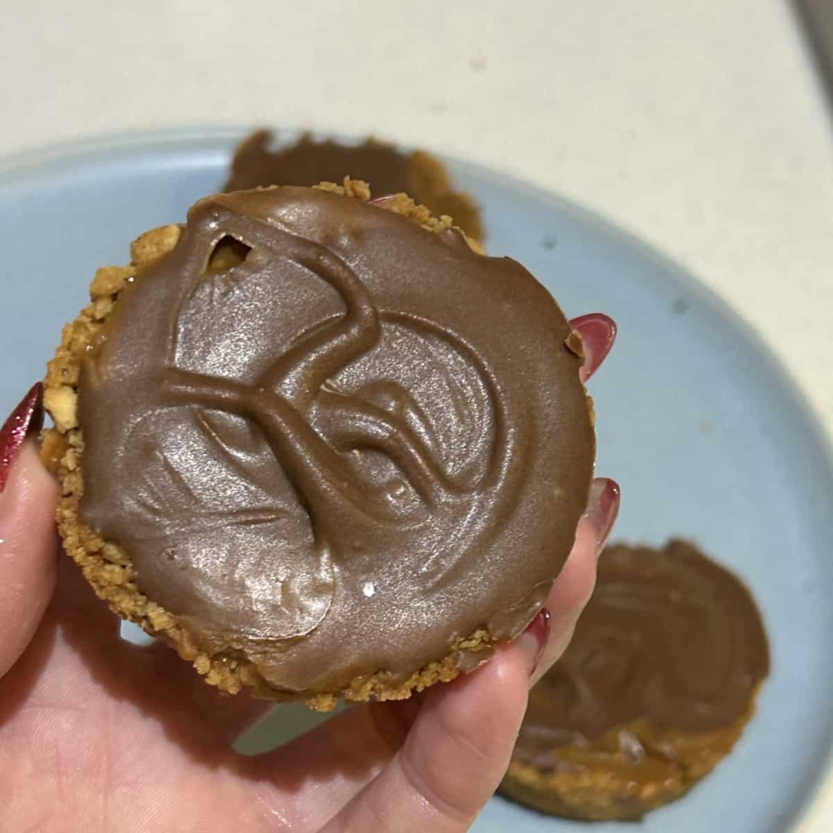 Foreground of image is a close up of a caramel tart being held up for the camera, background is caramel tarts sitting on a blue plate slightly blurred out.
