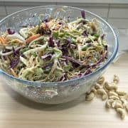 A side view of a large glass bowl containing a creamy noodle salad. Bowl is sitting on a light coloured chopping board decorated with some whole cashews.