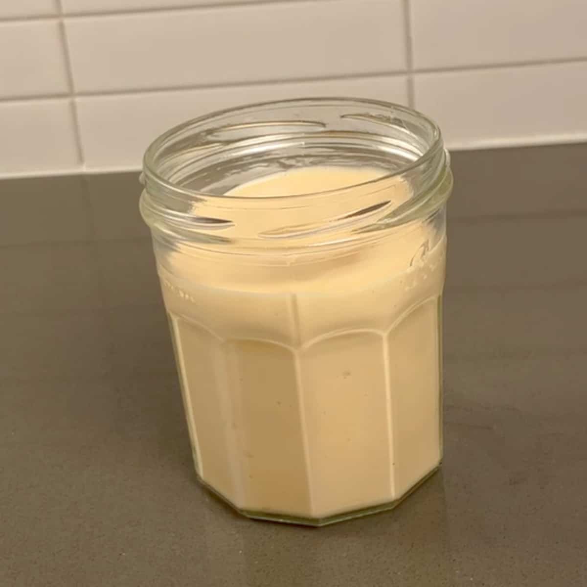 A glass jar with no lid containing mayonnaise sitting on a grey benchtop.
