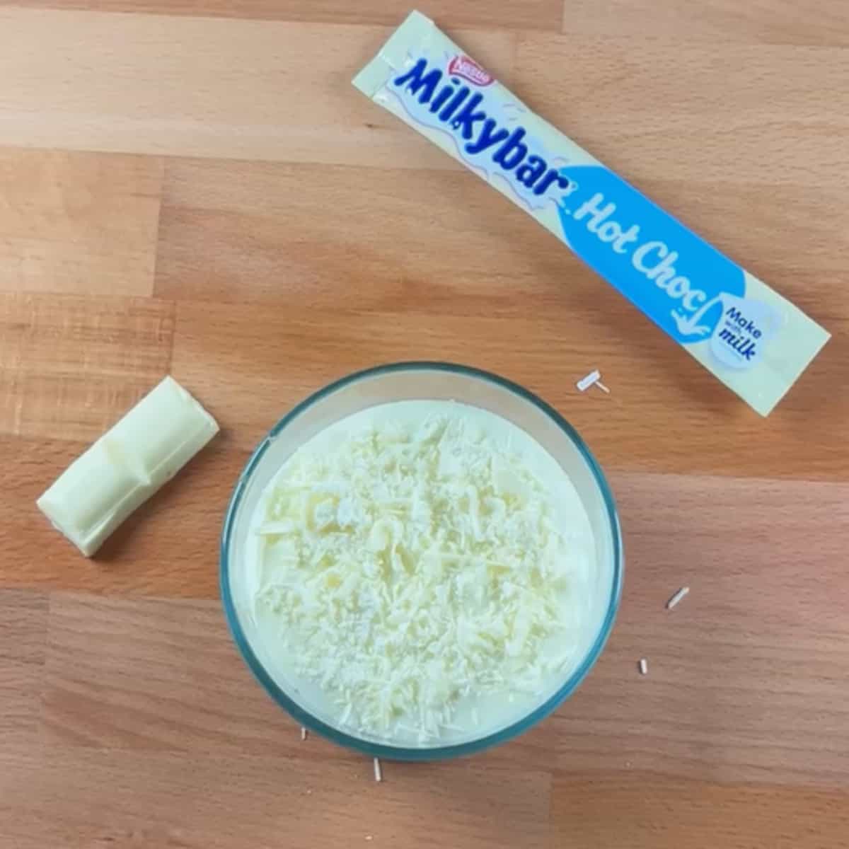 The centre of the image is a round food storage container containing Weet-bix soaked in milk and topped with yoghurt and shavings of white chocolate. A sachet of white-hot chocolate and two squares of white chocolate are placed to the side of the Weet-bix to decorate the image.